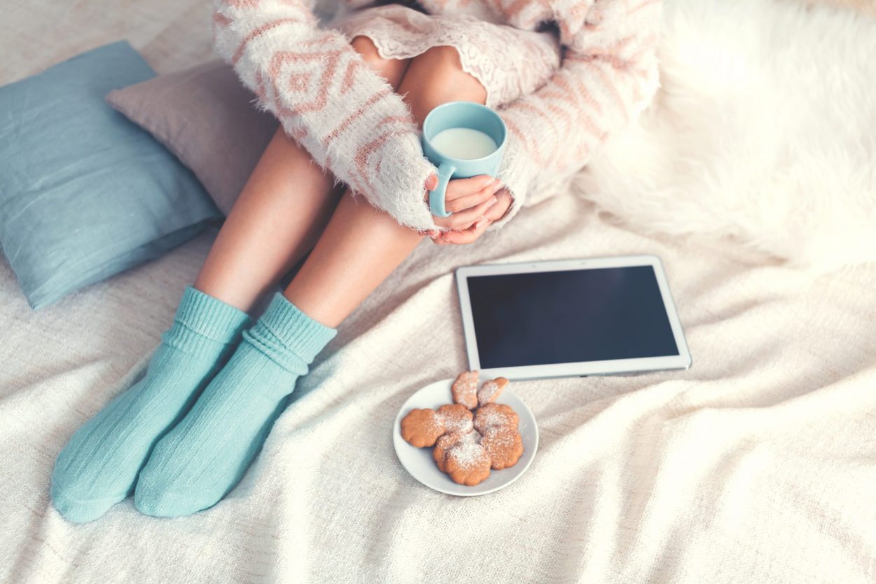 Woman sitting cozy on her bed in a sweater and socks, with her tablet, a mug of milk, and cookies.