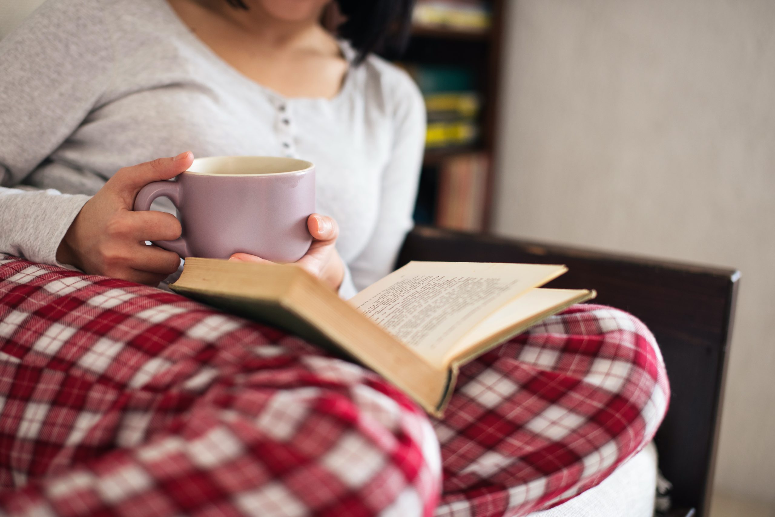 Close up of a woman's lap, she's wearing red buffalo check flannel pajama bottoms and a gray henley shirt, with an open book in her lap and a cup of coffee or tea, just a glimpse of her neck and short dark hair.